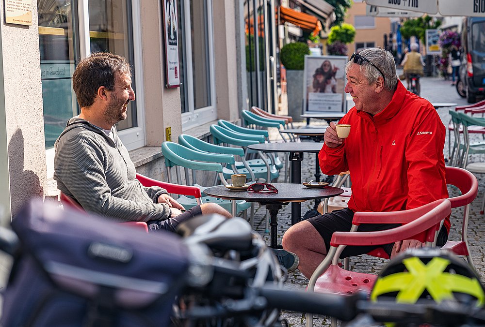 Zwei Männer unterhalten sich bei einem Espresso in einem Straßencafé. Im Vordergrund sind der Lenker eines Fahrrads und ein Fahrradhelm zu sehen Zwei Männer unterhalten sich bei einem Espresso in einem Straßencafé. Im Vordergrund sind der Lenker eines Fahrrads und ein Fahrradhelm zu sehen