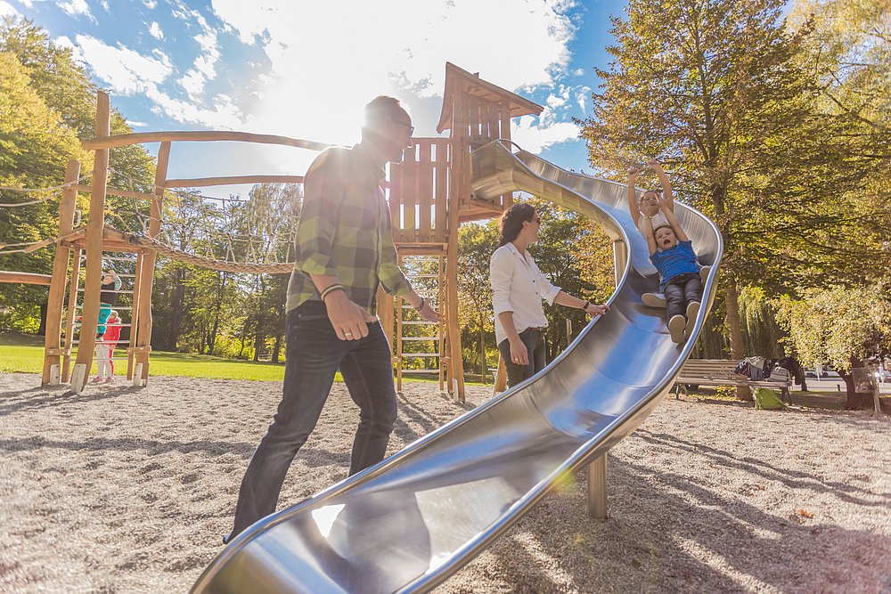 Eine Familie verbringt einen sonnigen Herbsttag im Klingelpark in Günzburg. Zwei Kinder rutschen lachend mit erhobenen Armen die Rutsche hinunter, während die Eltern danebenstehen. Eine Familie verbringt einen sonnigen Herbsttag im Klingelpark in Günzburg. Zwei Kinder rutschen lachend mit erhobenen Armen die Rutsche hinunter, während die Eltern danebenstehen.