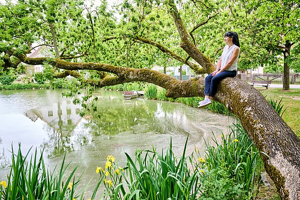 Junge Frau sitzt auf Ast eines Baumes, der über einen kleinen See ragt Junge Frau sitzt auf Ast eines Baumes, der über einen kleinen See ragt