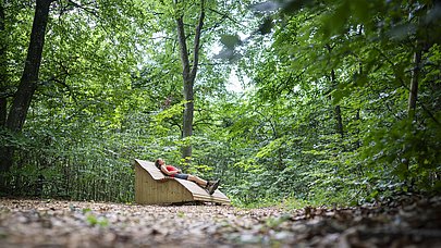 Eine Frau liegt auf einer großen Holzliege mitten im Wald und ruht aus