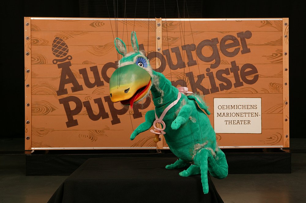 Die Kiste - Das Augsburger Puppenmuseum - Urmel vor dem Logo Die Kiste - Das Augsburger Puppenmuseum - Urmel vor dem Logo