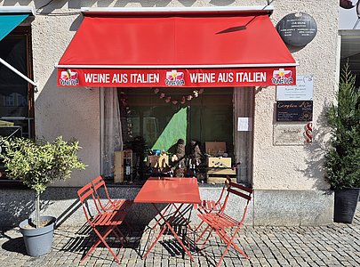 Eine rote Markise mit der Aufschrift ‚Weine aus Italien‘ hängt über einem Schaufenster. Davor stehen ein Bistrotisch und vier Stühle, die zu einer Pause bei Cappuccino einladen.“